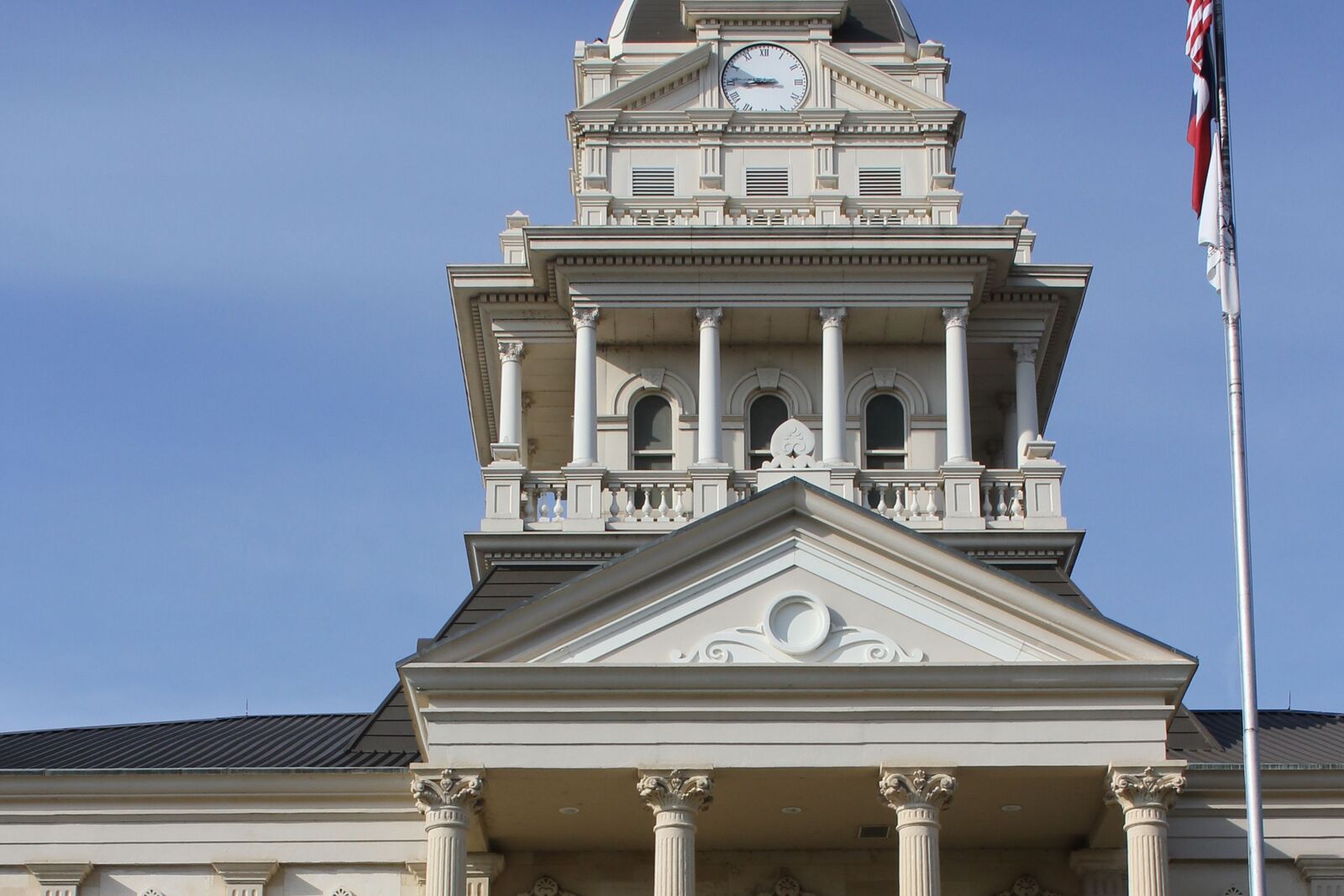 Clock tower, columned cupola and front pediment of a historic stone courthouse, with an American flag on a pole against a blue sky